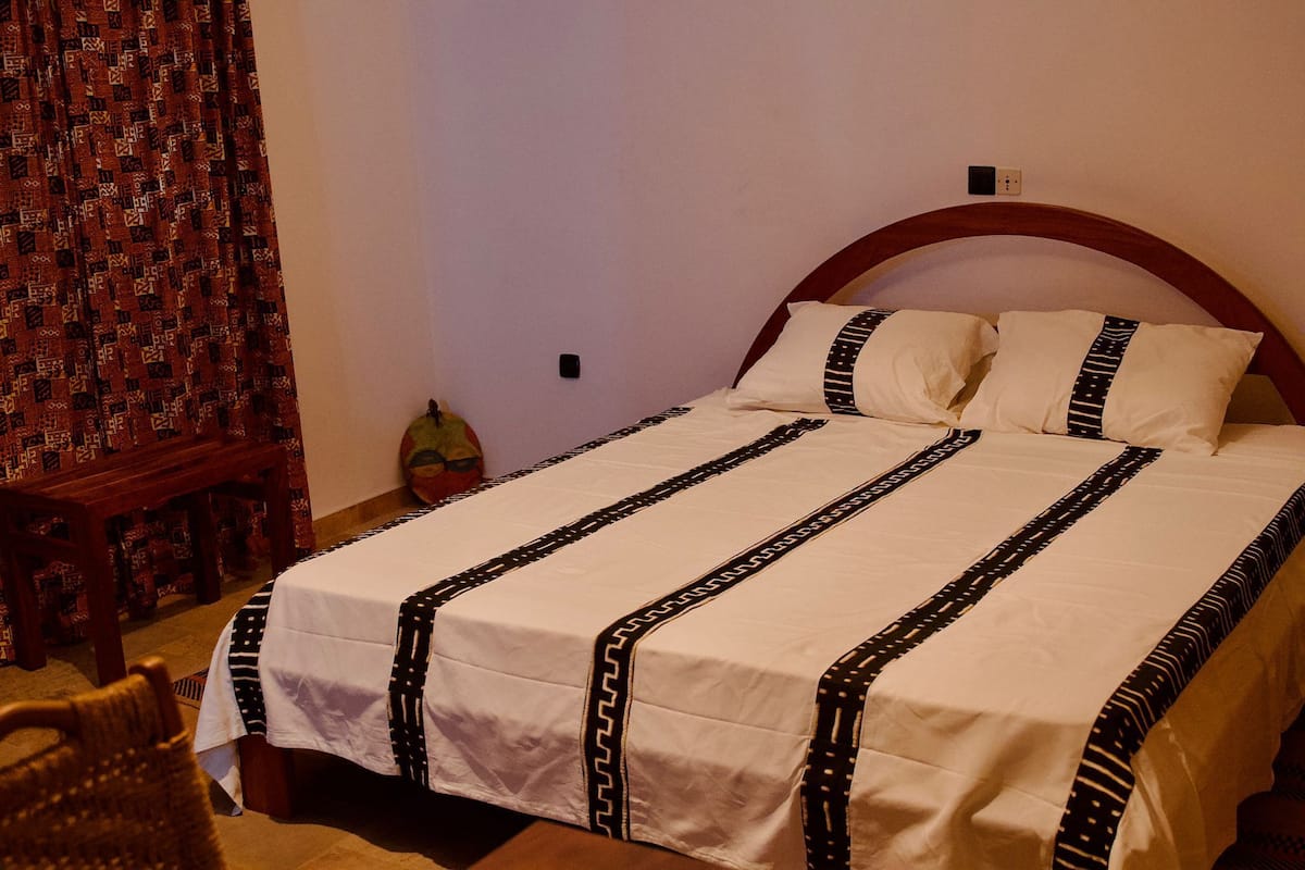 Cama bem arrumada com colcha estampada e almofadas, cabeceira de madeira e uma pequena cadeira de madeira.