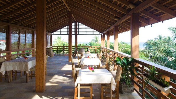 Un área de comedor al aire libre techada con mesas y sillas, vigas de madera y vistas al océano.