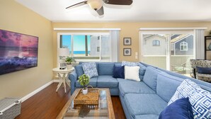 A living room with a blue sectional sofa, a ceiling fan, a large window with a view of the beach, and a painting on the wall. - 4 Bedroom Home Steps to the Sand in Newport Beach (Newport Beach)