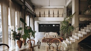 A spacious living area with a glass coffee table, wooden chairs, and large potted plants. A staircase with a metal railing leads to an upper level. - Mona Athens. (Athens)