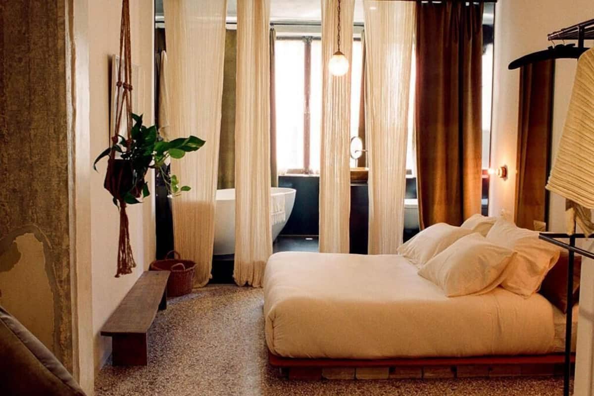 Ein Schlafzimmer mit einem Bett, Vorhängen, einer hängenden Pflanze, einer Bank und Blick ins Bad.