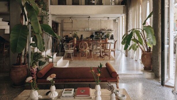 A spacious living area with a high ceiling, a brown sofa, multiple chairs, and large potted plants. - Mona Athens. (Athens)