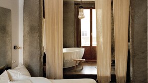 A bedroom with a large bed, a wooden nightstand, and a view of a bathroom with a bathtub. - Mona Athens. (Athens)