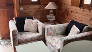 Interior acogedor de una cabaña con paredes de madera, un juego de sofá floral, una mesa blanca y una lámpara.