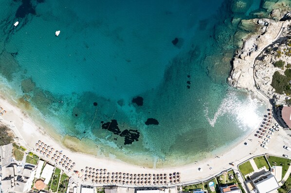 An aerial view of a beach with clear turquoise water, a sandy shore, and a row of umbrellas.