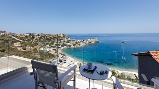Un balcón con una mesa, una silla y vistas a una zona costera con edificios y aguas azules cristalinas.
