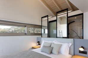 A modern bedroom with a large bed, a nightstand, a wall-mounted mirror, and a window with a view of a hilly landscape.