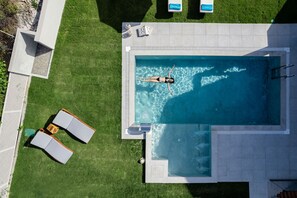 An aerial view of a swimming pool with a person in the water, surrounded by a well-maintained lawn and a patio area with lounge chairs.