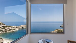 Habitación con una ventana grande que ofrece vista al mar, una pequeña mesa con una taza y un libro, y una silla.