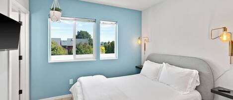 A bedroom with a bed, two windows, a hanging plant, a bedside table, and a wall-mounted light fixture.