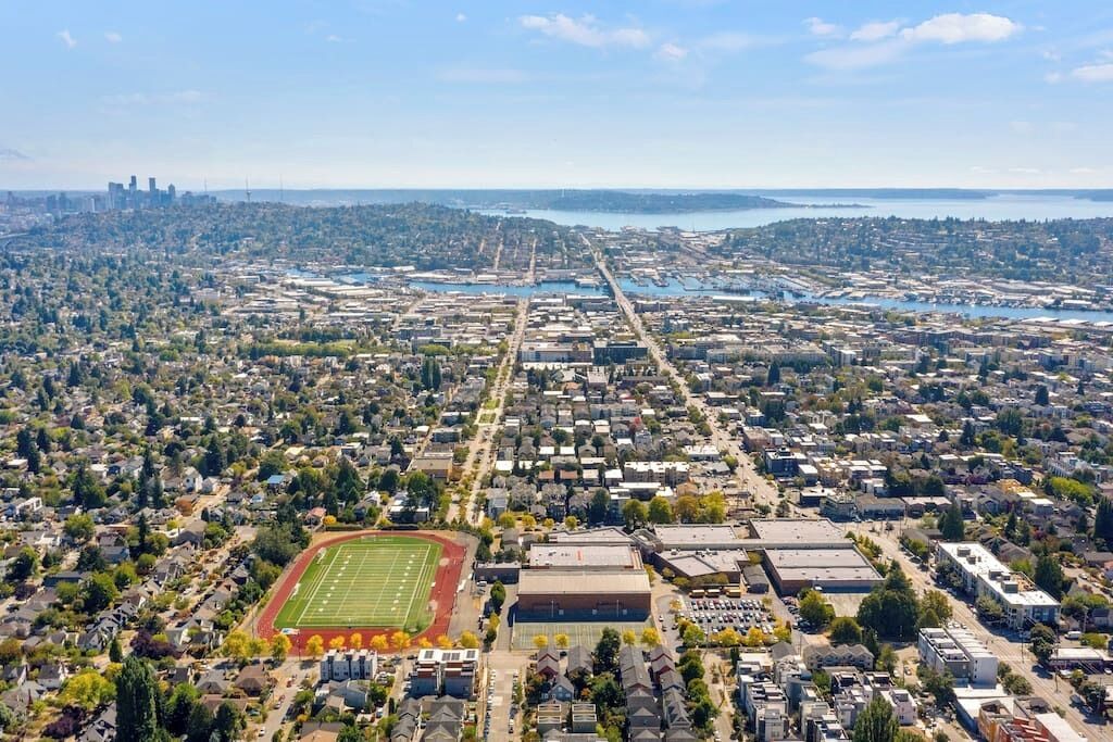 An aerial view of a cityscape with a large sports field, industrial buildings, and a waterfront.