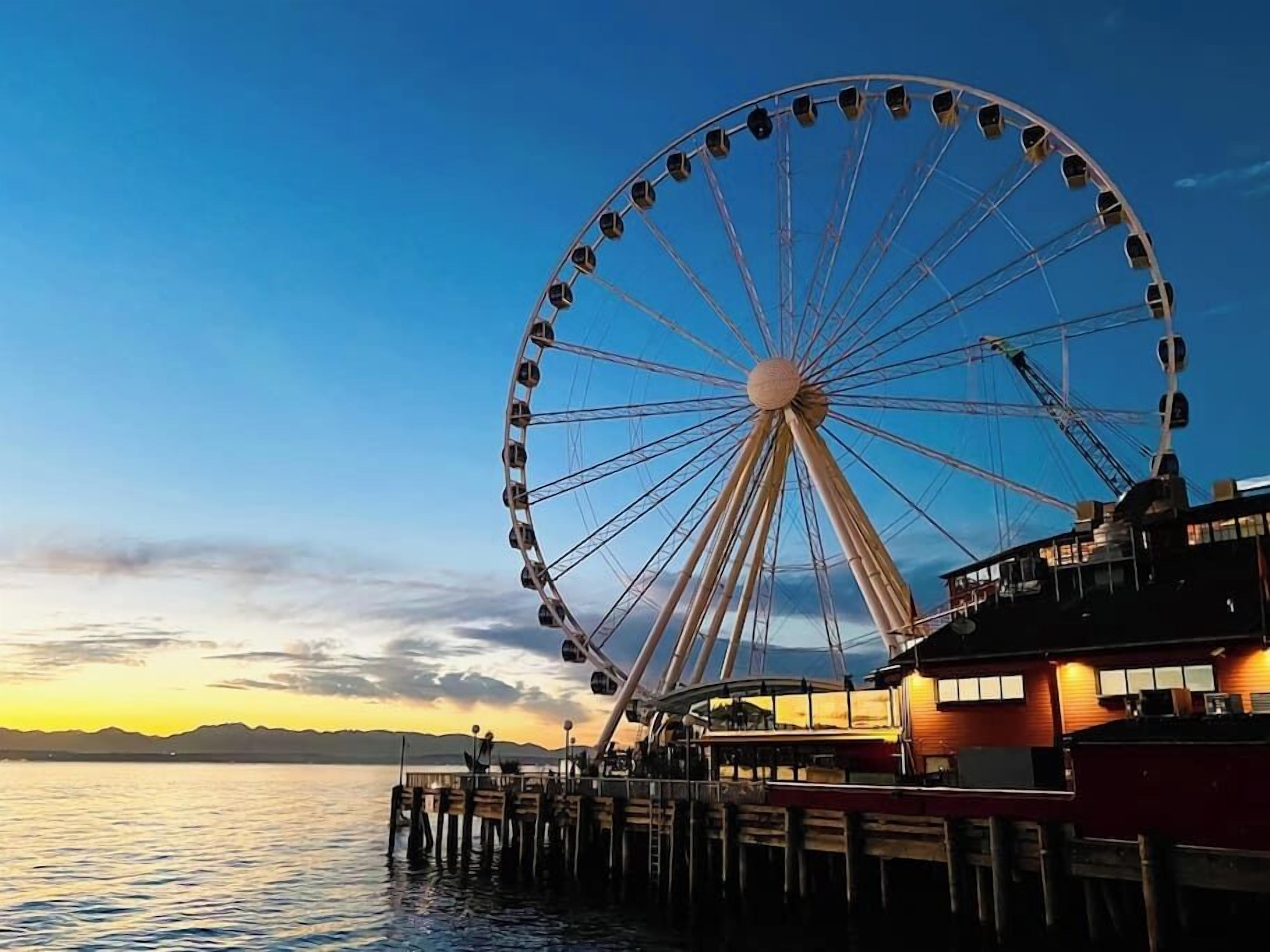 A Ferris wheel at dusk with a wooden pier extending into calm water, overlooking a distant shoreline.
