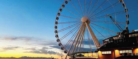 A Ferris wheel at dusk with a wooden pier extending into calm water, overlooking a distant shoreline.