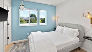 Un dormitorio con una cama, dos ventanas, una planta colgante, una mesita de noche y una lámpara fijada a la pared.