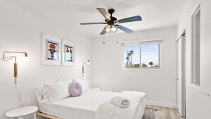A bedroom with a bed, a ceiling fan, a window, and artwork on the wall.