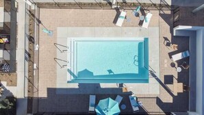 An aerial view of a rooftop pool area with a rectangular pool, lounge chairs, umbrellas, and a small seating area.