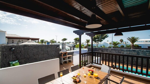An outdoor patio with a wooden table set for dining, a stone wall, and a view of a swimming pool. - 6 Bed Holiday Villa | Heated Pool | Hot Tub | Beach & Waterparks | Playa Blanca (Playa Blanca)