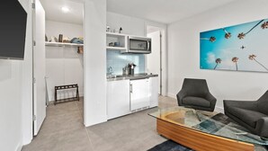 Un salon moderne avec une table basse en verre, deux fauteuils gris et un coin cuisine doté de meubles blancs et d’un pare-feu bleu.