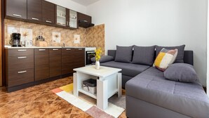 Ein modernes Wohnzimmer mit einem grauen Sofa, einem kleinen weißen Beistelltisch und einer Küchenzeile mit dunklen Holzschränken und einer gefliesten Spritzschutzwand.