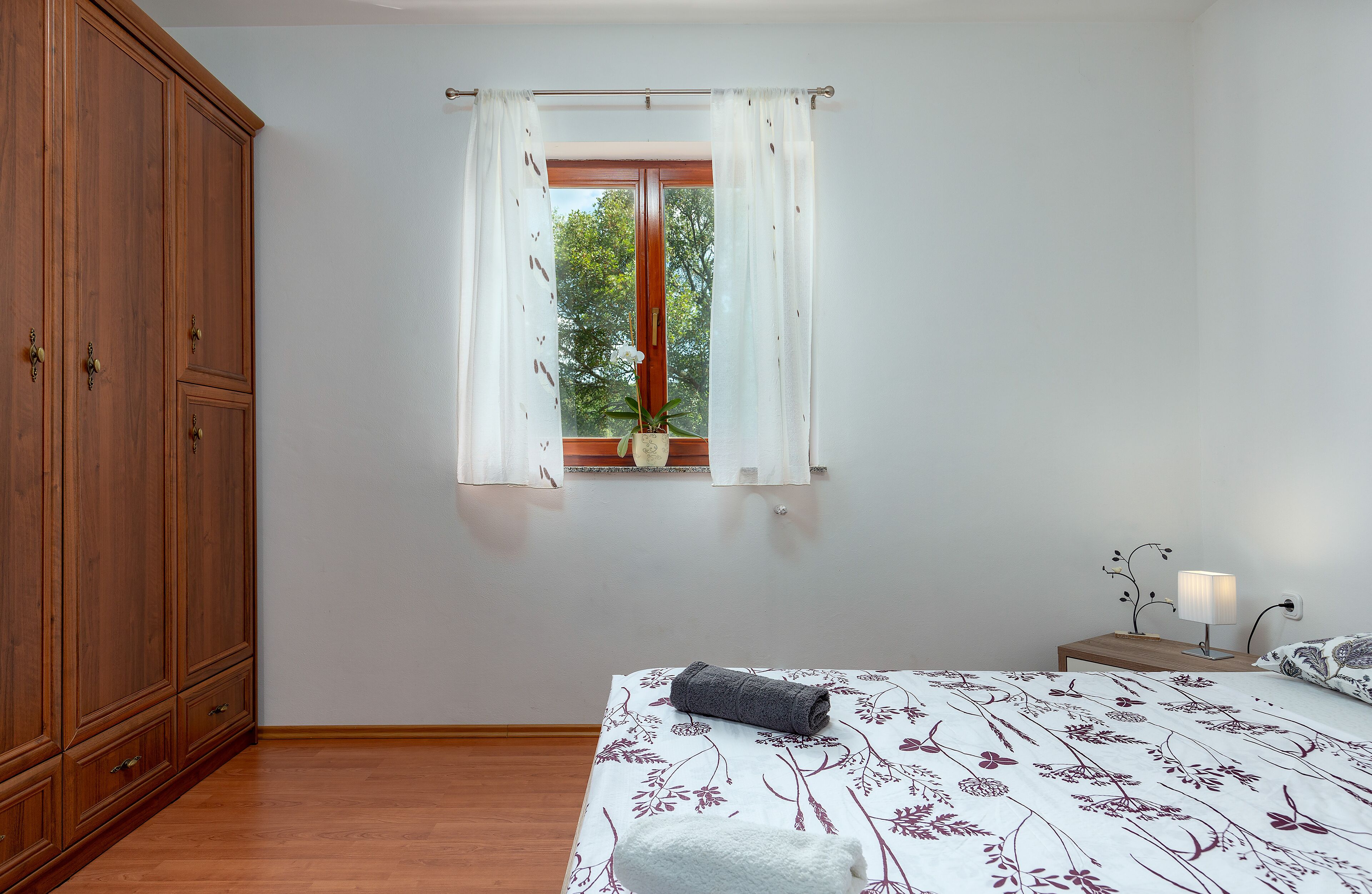 Een slaapkamer met een houten garderobe, een bed met bloemenbeddengoed, een raam met gordijnen en een nachtkastje met een lamp.