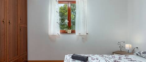 Een slaapkamer met een houten garderobe, een bed met bloemenbeddengoed, een raam met gordijnen en een nachtkastje met een lamp.