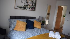 Ein Schlafzimmer mit Bett, Kissen, einem Nachttisch mit Lampe, einem Bild an der Wand und einem durchgehenden Tür erkennbaren Badezimmer.