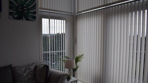 Ein Wohnzimmer mit einem grauen Sofa, einem kleinen Beistelltisch mit einer Lampe und einem Fenster mit vertikalen Jalousien.