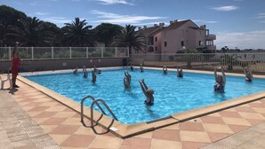 Une piscine où se trouvent plusieurs personnes, une terrasse carrelée et un bâtiment en arrière-plan.