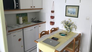 Une cuisine compacte équipée d’un four à micro-ondes, d’un évier et d’un petit réfrigérateur. Une table à manger avec des chaises est placée devant la cuisine.