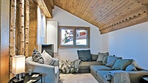 Un salón acogedor con techo de madera alto, una chimenea y vistas a un pueblo nevado a través de una ventana.