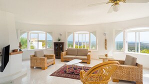 Un salón amplio con sofá, sillas, televisor y ventanas grandes que ofrecen vistas del paisaje.