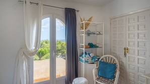 Una habitación con una puerta blanca, una silla blanca con un cojín azul, un estante blanco con toallas dobladas y vistas a través de una ventana con cortina.