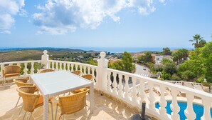 Una terraza con mobiliario de mimbre, barandilla blanca y vistas al mar y al paisaje.