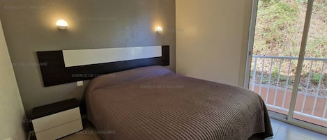 Un dormitorio con una cama grande, una mesita de noche, una lámpara de pared y vistas al exterior a través de un ventanal corredero.