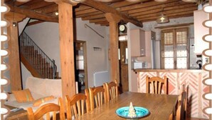 Uma sala de jantar com mesa e cadeiras de madeira, um relógio na parede e cozinha ao fundo.