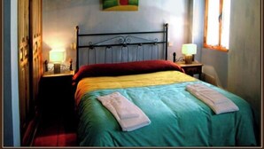 Um quarto com estrutura de cama de metal, colcha verde, travesseiro vermelho e um quadro de uma paisagem na parede.