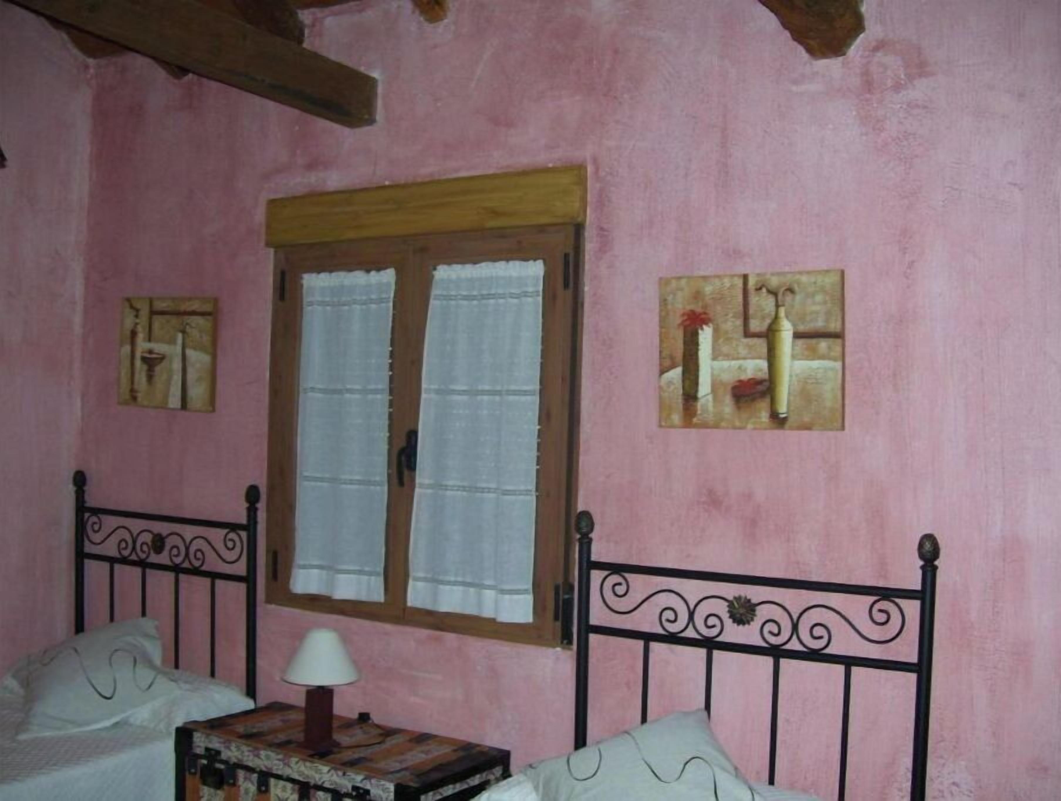 A room with a bed, a small table with a lamp, a window with curtains, and two wall-mounted paintings.