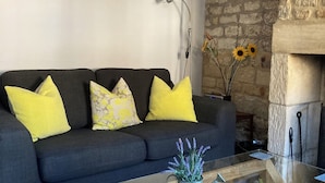 Un salon moderne avec un canapé gris foncé, des coussins jaunes et à motifs, une table basse en verre surmontée d’un vase en métal et une cheminée en pierre.