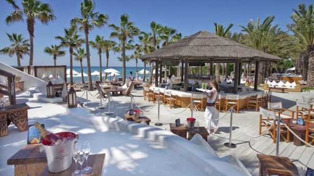 Een strandresort met terras, palmbomen en een paviljoen met rieten dak.