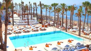 Een zwembadterras met ligbedden en parasols, palmbomen en uitzicht op de oceaan.