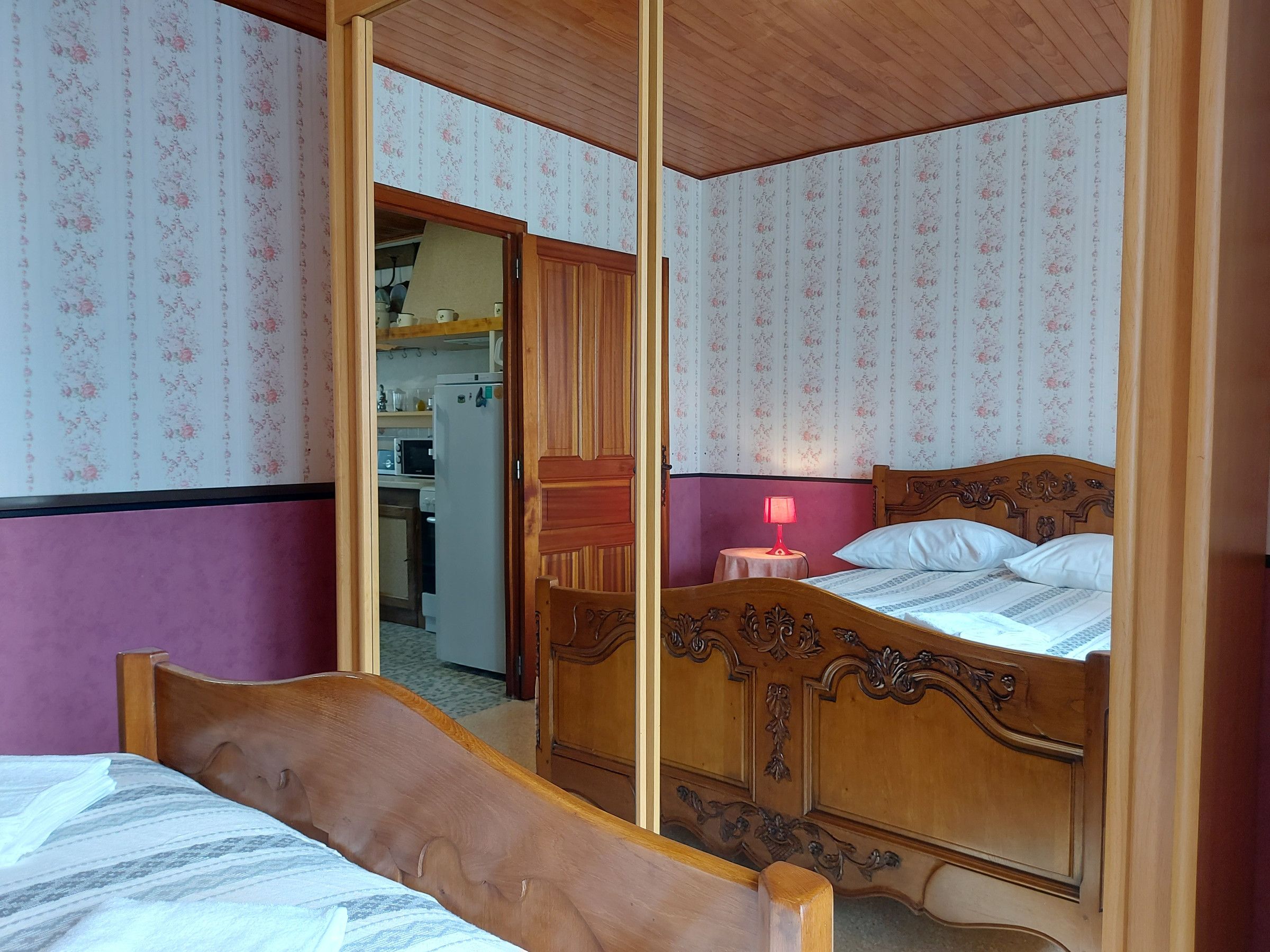 A bedroom with a wooden bed, a small bedside table with a lamp, and a view into the kitchen.