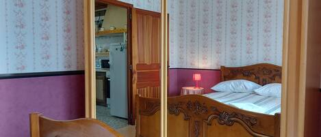 A bedroom with a wooden bed, a small bedside table with a lamp, and a view into the kitchen.