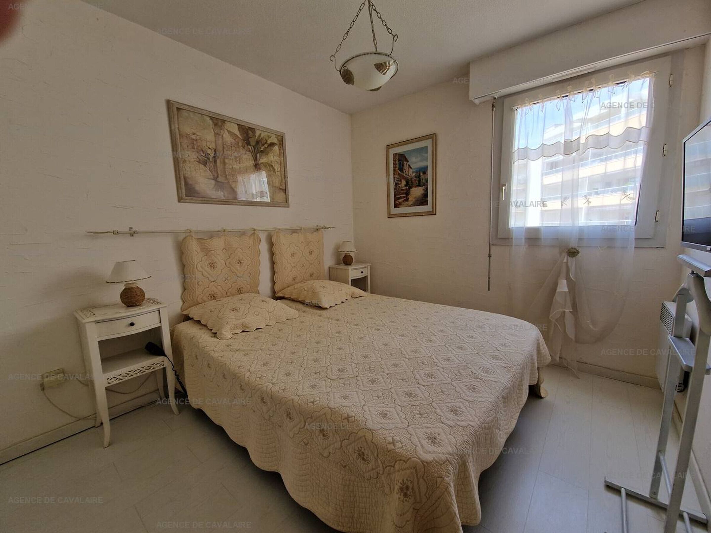 Ein Schlafzimmer mit einem großen Bett, einem Nachttisch mit Lampe, einem kleinen Tisch mit Radio und einem Fenster mit Vorhängen.