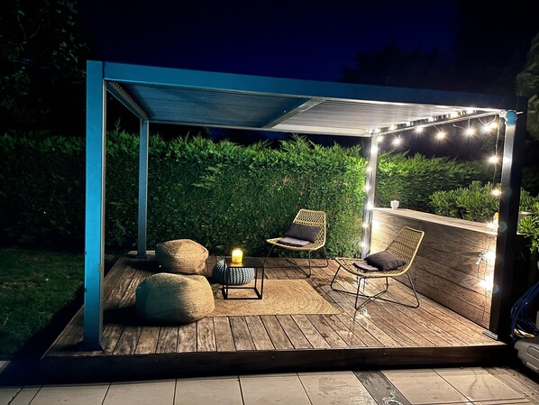 An outdoor patio with wooden flooring, two wicker chairs, a small table with a candle, and string lights.