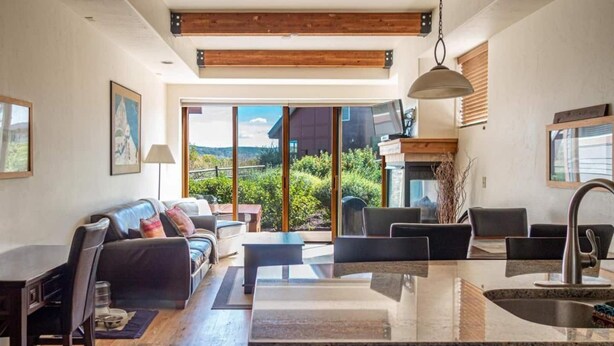 A modern kitchen with a central island, a dining area with a sofa and chairs, and a large sliding glass door leading to a scenic outdoor view. - Great Rates, Hot Tub, Tranquil Views & Location (Park City)