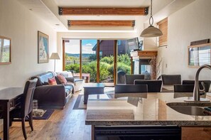 A modern kitchen with a central island, a dining area with a sofa and chairs, and a large sliding glass door leading to a scenic outdoor view. - Great Rates, Hot Tub, Tranquil Views & Location (Park City)