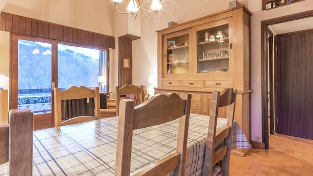 A dining area with a wooden table and chairs, a large cabinet with glass doors, and a view of a mountainous landscape through a large window. - Charming 3-room with mezzanine for 6 pers, south balcony and parking in La Clusaz (La Clusaz)