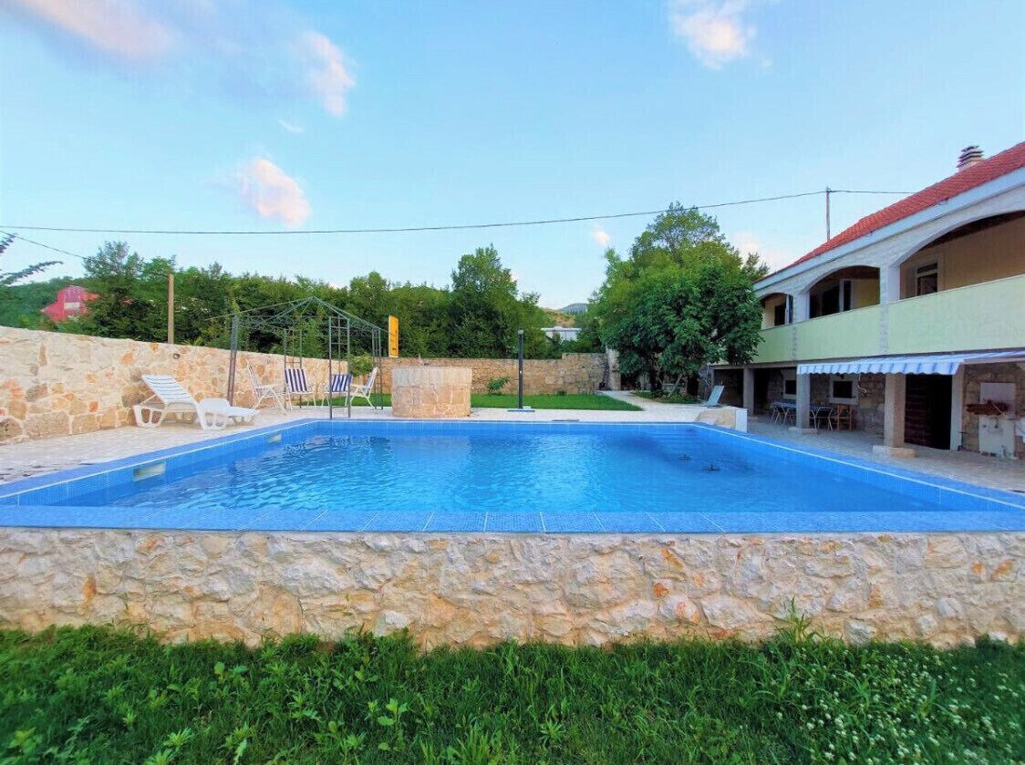 Una piscina con paredes de piedra, rodeada de vegetación y un edificio con techo rojo.