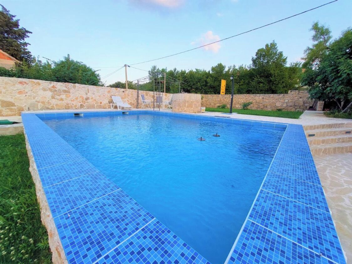 A rectangular swimming pool with blue mosaic tiles, surrounded by a stone wall and a grassy area.