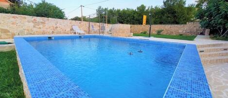 A rectangular swimming pool with blue mosaic tiles, surrounded by a stone wall and a grassy area.